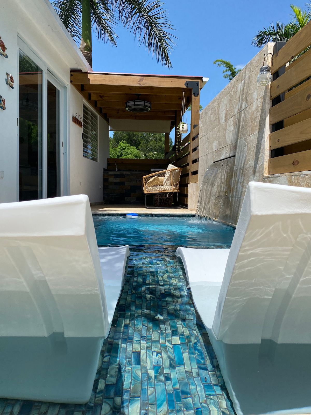 Two white lounge chairs are placed partially submerged in the crystal-clear water of the private pool. A shaded seating area with a wicker chair is visible in the background, surrounded by wooden fencing and tropical vegetation, under a clear blue sky.