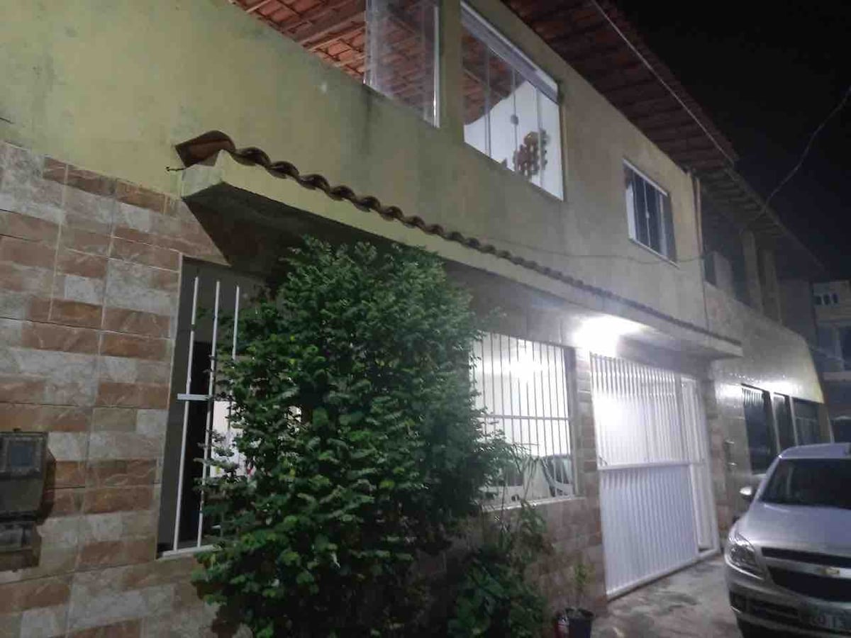 The exterior of a two-story accommodation is showcased at night, with warm light emanating from the windows. A bush provides greenery near the entrance, while a vehicle is parked in front. The building features a tiled roof and textured wall surfaces.
