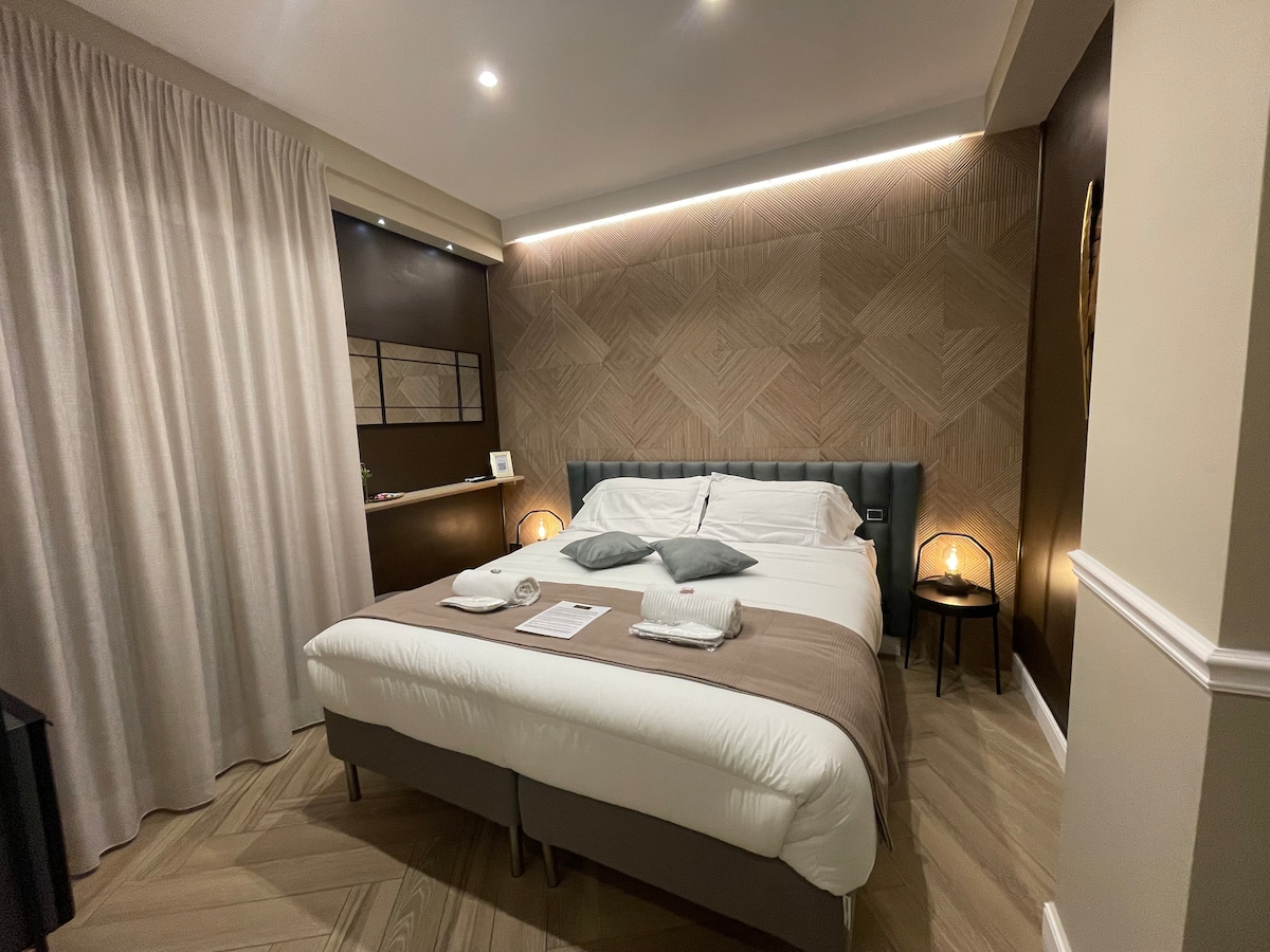 A modern bedroom features a spacious bed with plush bedding and decorative pillows. Soft lighting is provided by bedside lamps. The walls are adorned with contemporary design elements, and a textured pattern adds visual interest. A curtain softly drapes over a window, complementing the elegant decor.