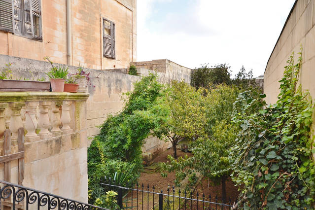 A serene garden area is visible, surrounded by stone walls. Lush greenery fills the space, including trees and plants along the pathways. A decorative railing adds character, while a glimpse of a historic building is seen in the background, contributing to the quaint atmosphere.