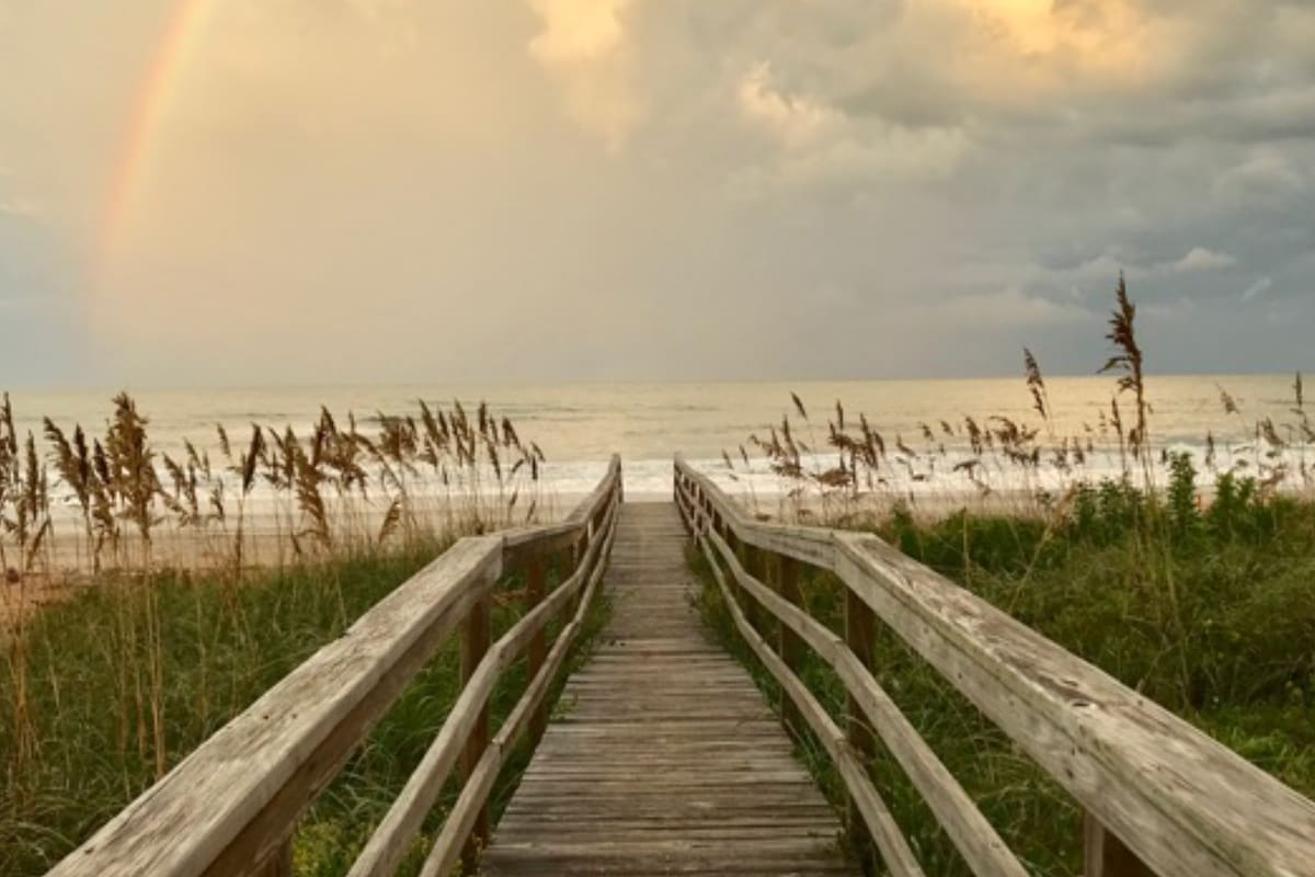 A wooden boardwalk leads through tall grass toward the ocean. Soft waves are visible under a cloudy sky, with hints of a rainbow appearing above. The scene captures a tranquil coastal atmosphere, inviting exploration of the beachfront.