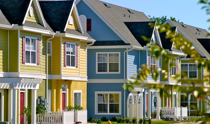 A row of colorful two-story residences is presented, featuring a variety of hues including yellow, blue, and light green. Each home showcases large windows and distinctive architectural details. Lush greenery and decorative pathways accentuate the inviting atmosphere of this vibrant neighborhood.