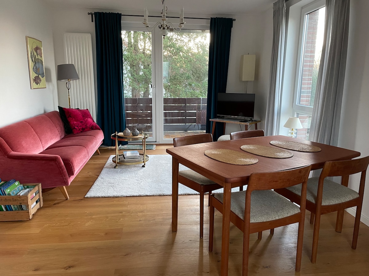 A living area features a pink sofa with decorative pillows and a wooden dining table surrounded by four chairs. A light rug covers the wooden floor, while a television sits on a stand. Windows offer natural light and views of the balcony.