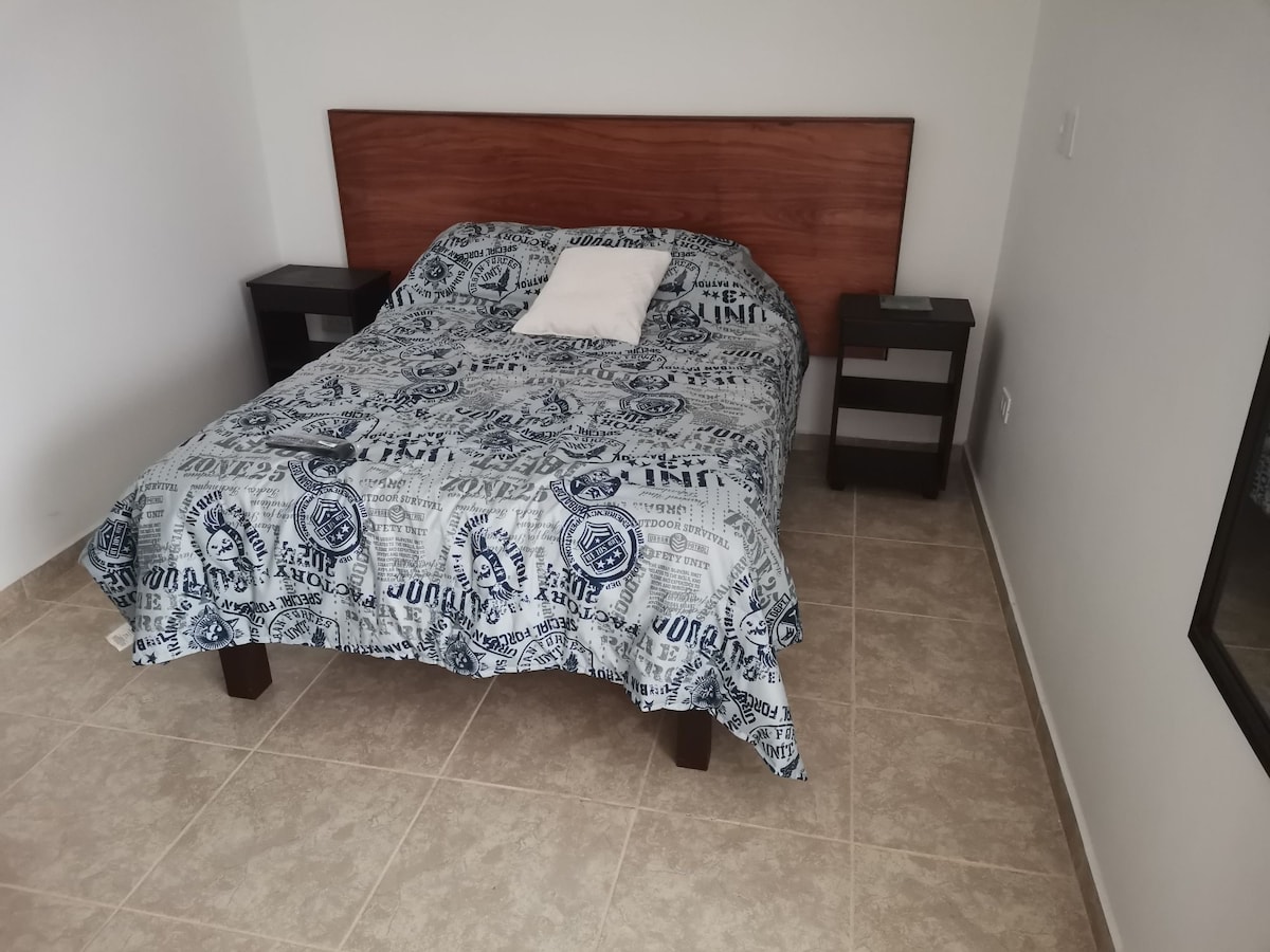 A bedroom features a matrimonial bed covered with a patterned blanket. Nightstands are positioned on either side of the bed. The room is illuminated by natural light, highlighting the neutral-colored walls and tiled flooring.