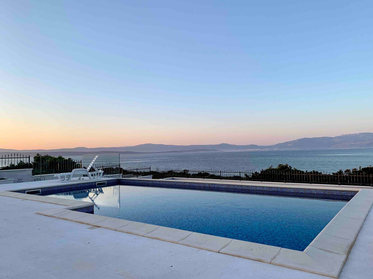 A serene outdoor pool is positioned elegantly against a backdrop of the sea at sunset. The waters reflect soft hues of blue and orange, with a spacious deck surrounding the pool. A white lounge chair is visible nearby, enhancing the tranquil ambiance.