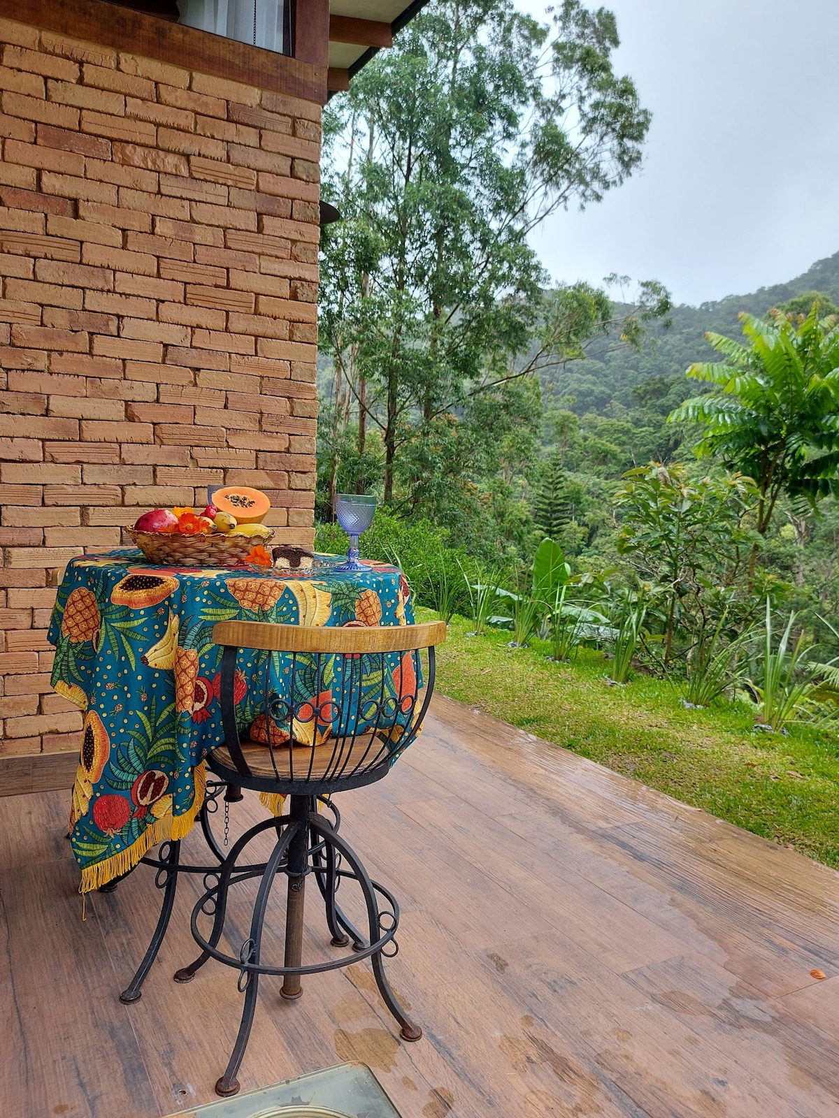 A small outdoor dining area is displayed, featuring a round table adorned with a colorful tablecloth and a bowl of fresh fruit. A barstool provides seating, while lush greenery and distant mountains are visible in the background, creating a peaceful natural setting.