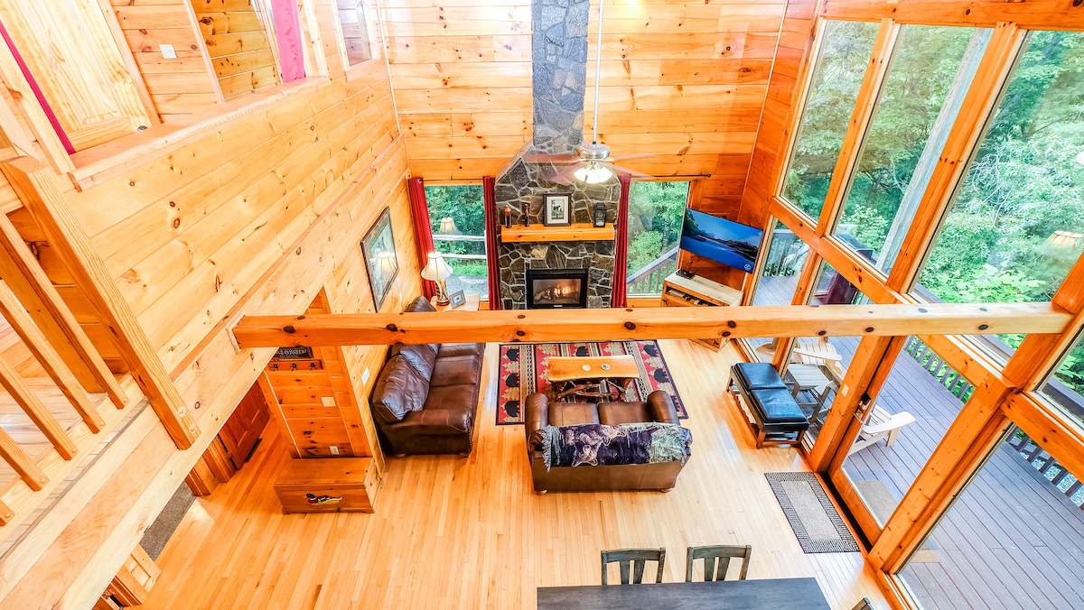 An aerial view of the Great Room showcases high, wooden ceilings and a wall of windows that capture natural light. A stone fireplace is positioned below, surrounded by comfortable seating. Rustic decor accents the space, with a wooden dining table visible in the foreground.