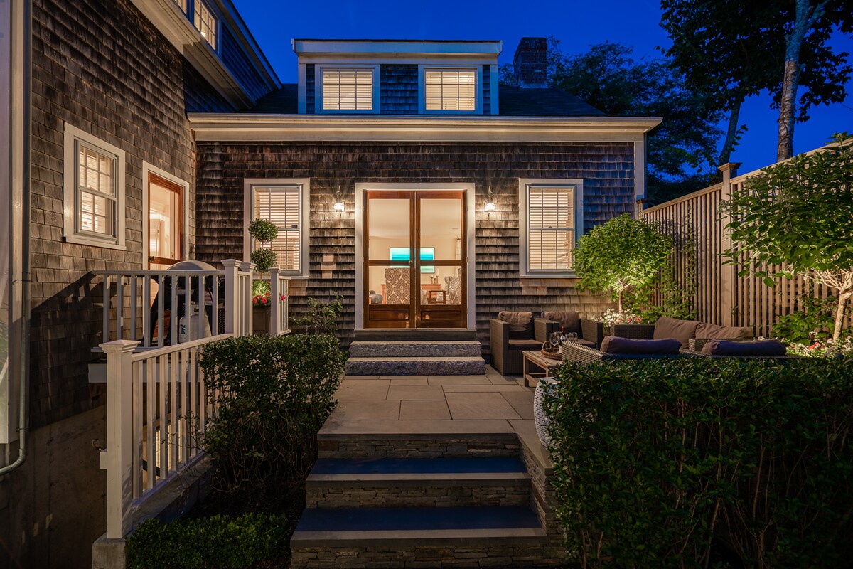 The exterior of the house is shown at dusk, with inviting light spilling from the windows. Steps lead up to a small patio, surrounded by lush greenery. A seating area with comfortable furniture is visible, providing a welcoming atmosphere for relaxation.