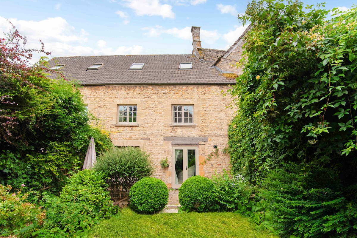 The exterior of the stone cottage is framed by lush greenery, highlighting its rustic charm. A well-maintained garden with manicured bushes leads to a pair of double doors, inviting entry to the interior. The structure features a slate roof and large windows set into the walls.