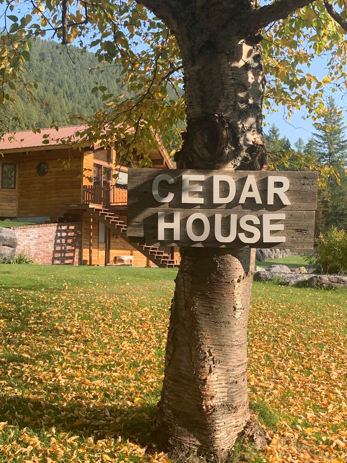 A wooden sign reading 'CEDAR HOUSE' is prominently displayed on a tree, with a backdrop of a rustic wooden home and a landscape of falling autumn leaves. The home features a staircase leading to an elevated entryway amidst lush greenery.