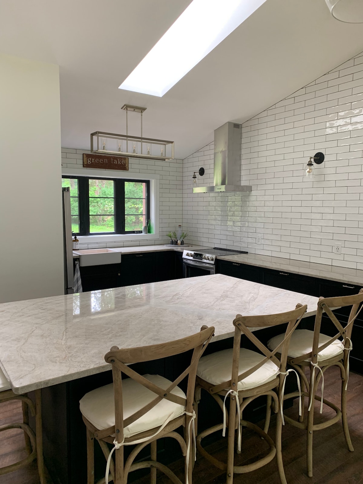 A modern kitchen features a large island with seating for four, complemented by a sleek marble countertop. The space is illuminated by a skylight and adorned with white subway tiles. Windows provide views of the surrounding greenery, enhancing the ambiance.