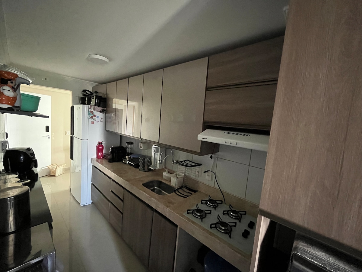 A functional kitchen is equipped with wooden cabinetry and a countertop made of light-colored stone. Essential appliances include a gas stove and refrigerator. Shelves hold various kitchenware, while ample counter space is available for meal preparation.