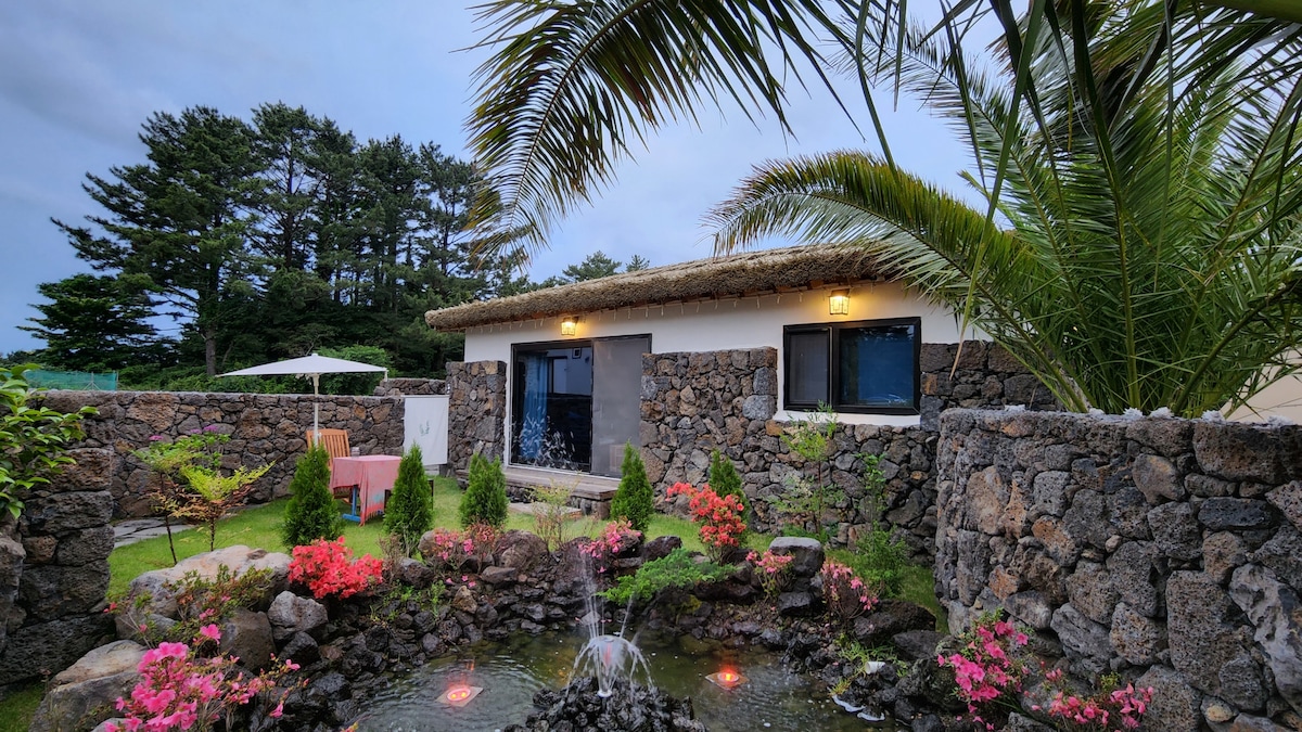 A serene outdoor space features a stone wall surrounding a well-maintained garden. Colorful flowers add vibrancy, while a small water feature gently bubbles in the center. The building showcases large windows and a thatched roof, enhancing the natural ambiance of the setting.
