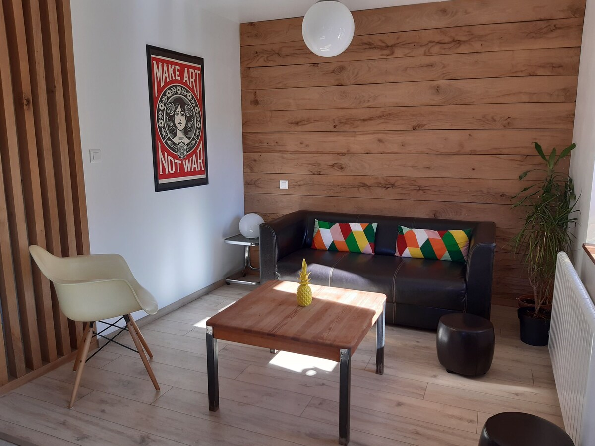 The inviting living area features a dark leather sofa adorned with colorful cushions. A wooden coffee table is positioned centrally, accompanied by a small round stool. The wall behind the sofa is panelled with warm wood, and a vibrant art piece adds character to the space.