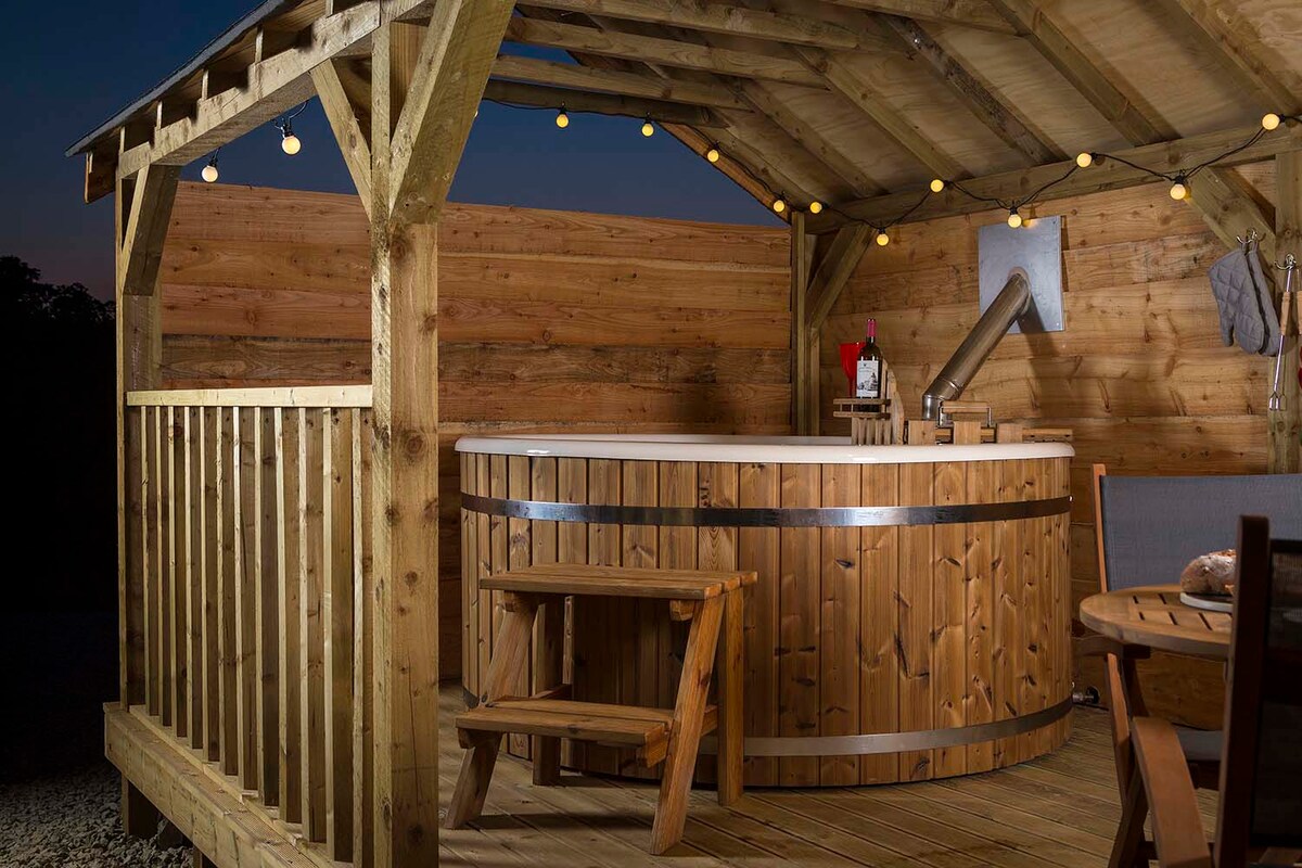 A wooden structure features a round wood-fired hot tub under a covered area. String lights illuminate the space, creating a cozy ambiance. A wooden bench and round table are placed nearby, offering a lovely spot to relax by the hot tub.