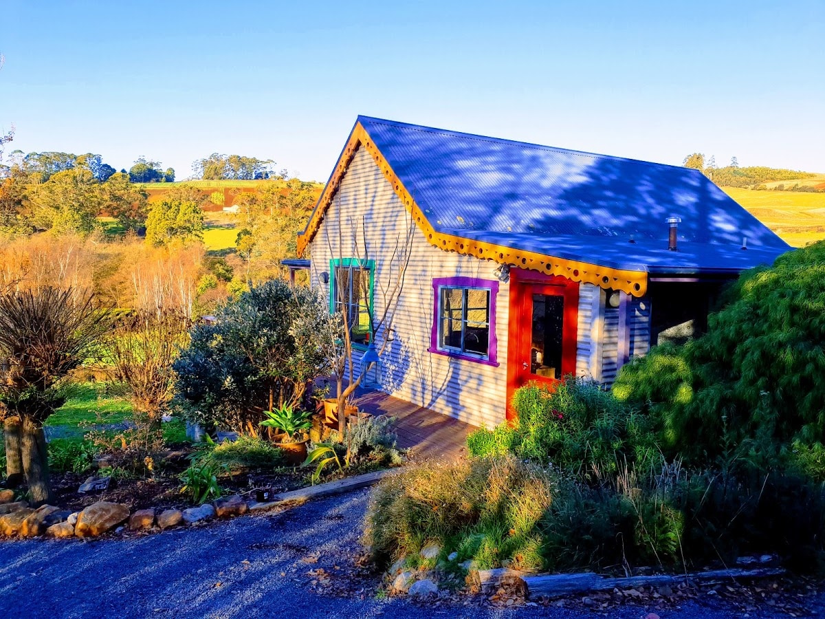 A charming cottage is nestled among rolling hills, featuring a colorful façade with a blue roof and vibrant door. Surrounding gardens are filled with various plants and shrubs, adding to the natural setting. A gravel path leads to the entrance, showcasing a peaceful landscape.