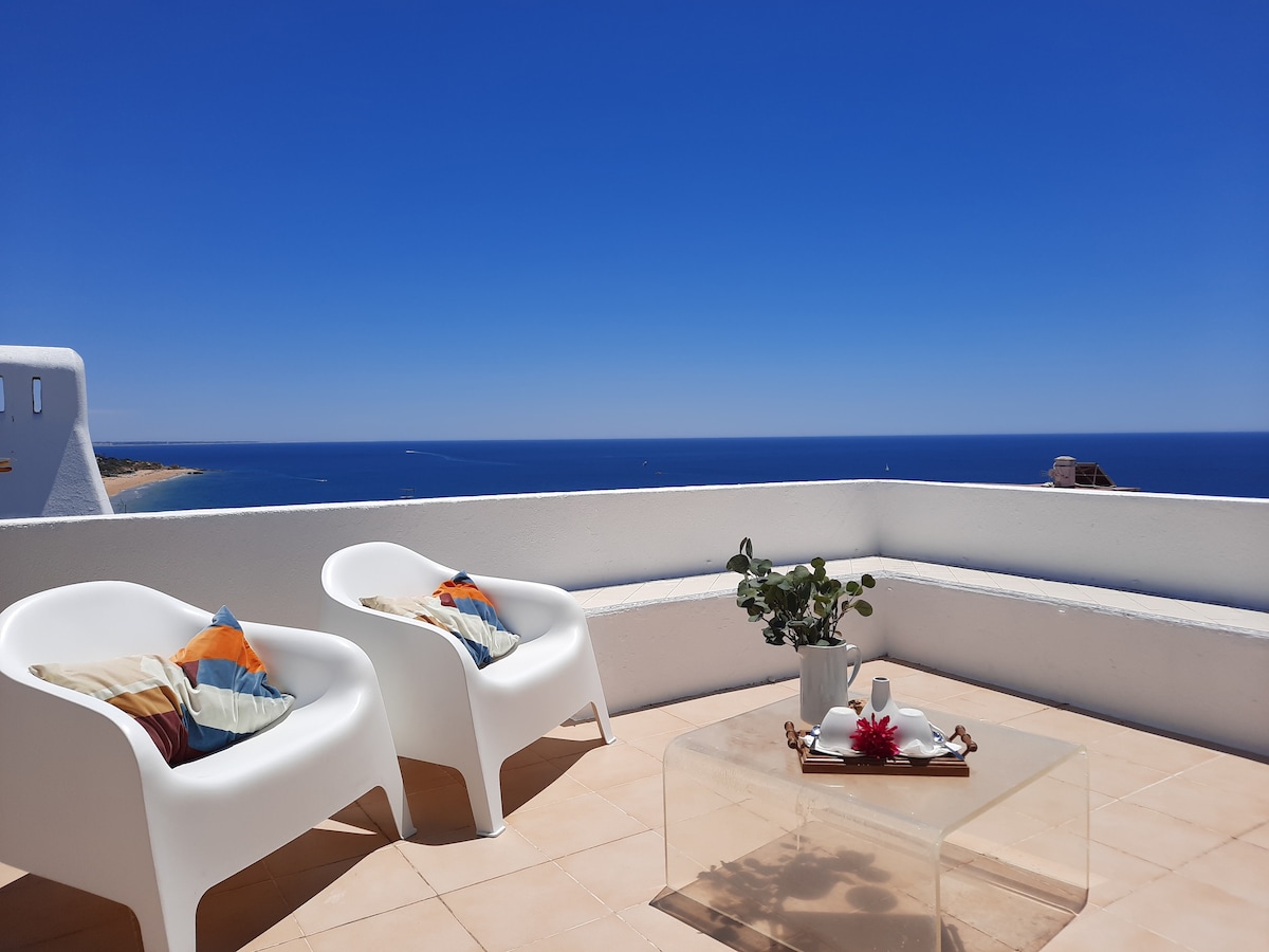 A spacious terrace offers a breathtaking view of the ocean, with two contemporary white chairs adorned with decorative cushions. A glass table is positioned between them, featuring a small potted plant and tasteful decor, creating a serene setting for relaxation.
