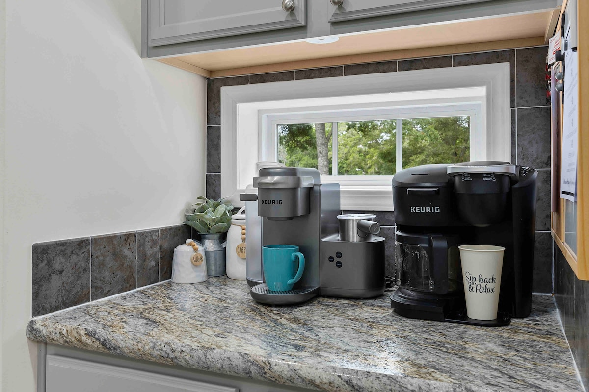 A kitchen corner is presented with a Keurig coffee maker and an array of accessories on a granite countertop. A blue mug rests beside a silver brewing machine, with an assortment of canisters nearby. Natural light enters through a small window, enhancing the inviting space.