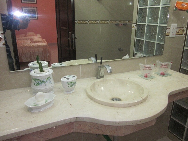 A bathroom countertop is visible, featuring a cream-colored sink surrounded by a curved marble surface. Decorative items, including a small plant and toiletries, rest on the counter. A mirror reflects the room's light, providing a bright ambiance. The background shows the entrance to a bedroom.