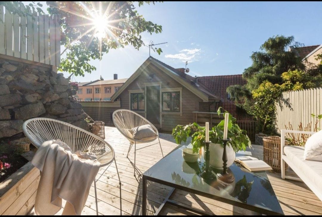 A sunlit deck area features two stylish chairs alongside a table topped with decorative items. Lush greenery frames the space, while a cozy blanket is draped over one of the chairs. Mountain views can be seen in the distance, enhancing the inviting outdoor setting.