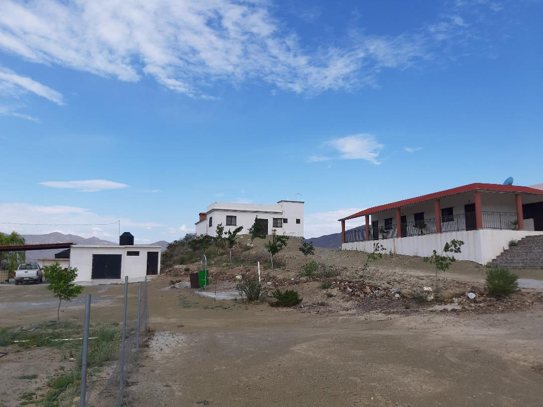 A ranch-style property is visible, featuring two separate buildings under a clear blue sky. The main structure is white with dark trim, while the adjacent building has a red-tiled roof. Surrounding terrain consists of gravel and sparse vegetation, providing a natural setting.