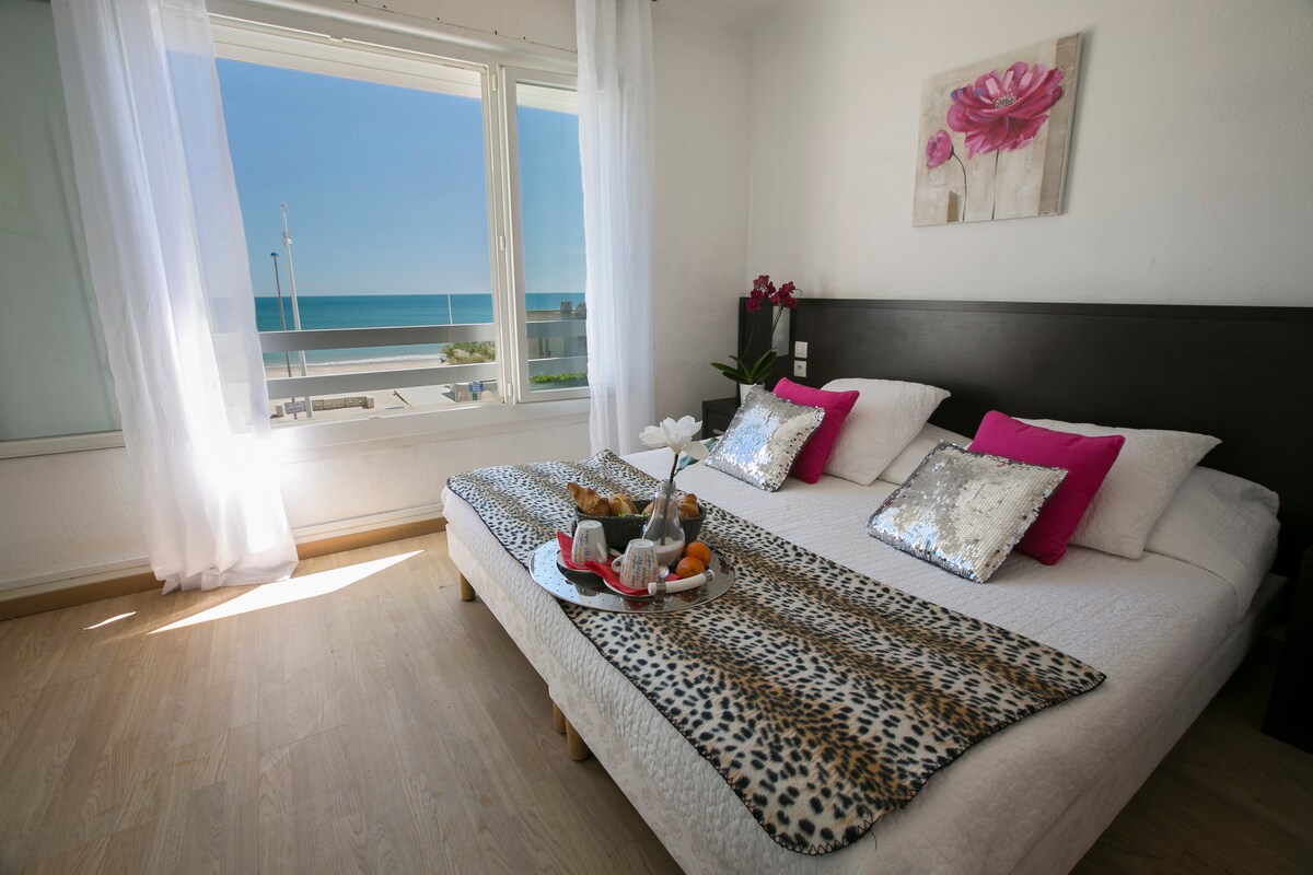 A bright room features a spacious bed adorned with decorative pillows and a stylish throw. A tray with breakfast items rests on the bed. Large windows allow natural light to illuminate the space and offer a stunning view of the sea and beach.