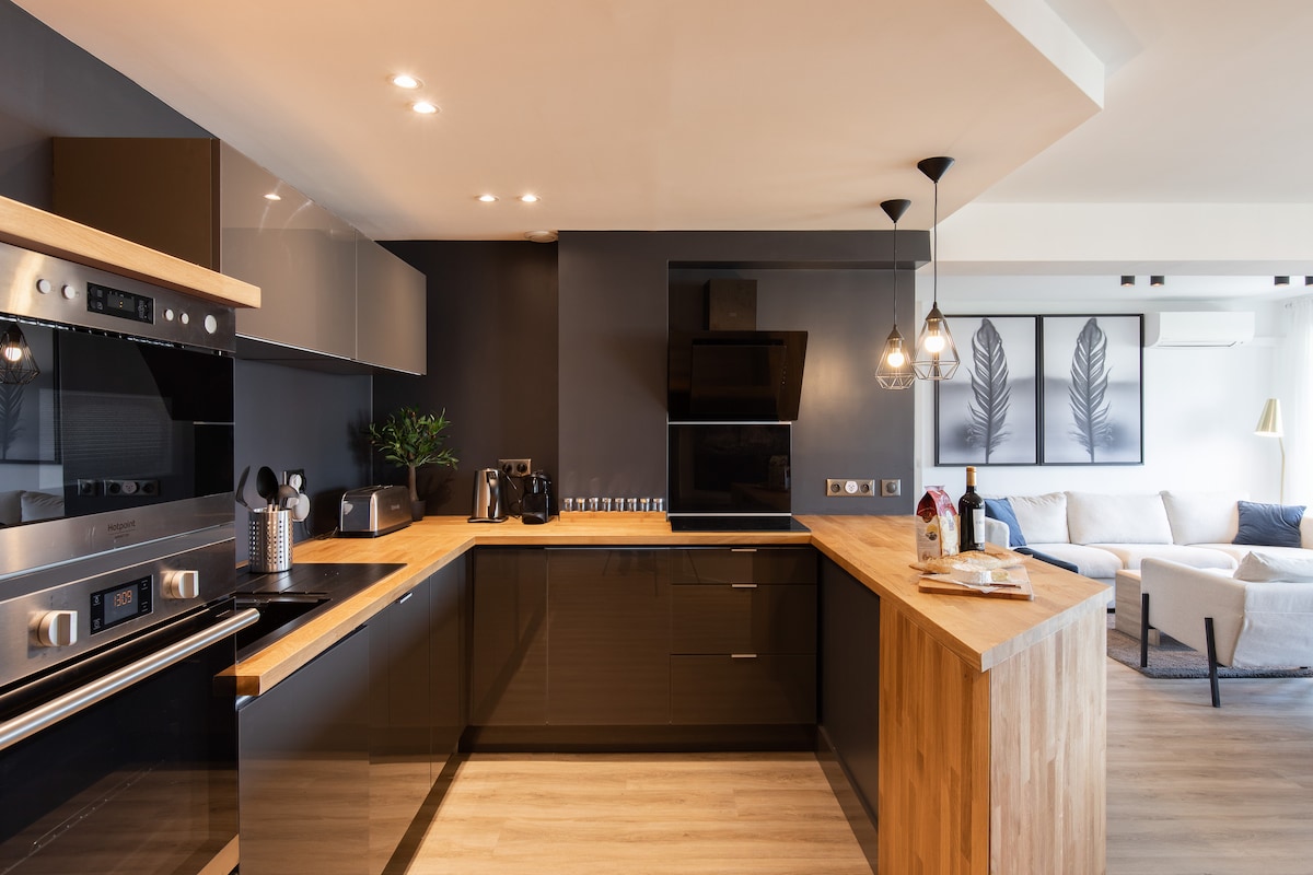 A modern kitchen features sleek black cabinetry paired with a warm wood countertop. High-end appliances, including an oven and coffee machine, are visible. Pendant lights add subtle illumination, creating a practical yet inviting cooking space adjacent to a softly furnished living area.