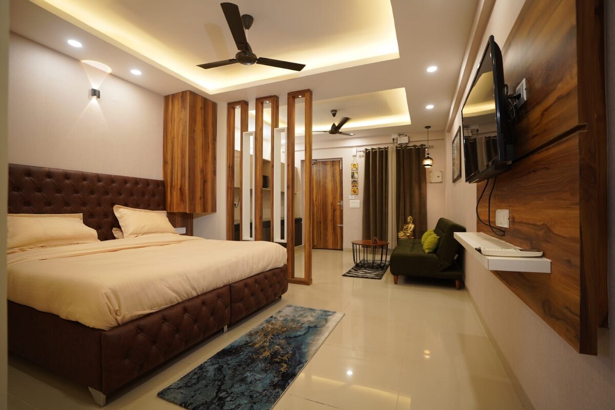 A spacious bedroom is showcased featuring a large, tufted bed dressed in neutral-toned linens. Light wood accents complement the modern design, while a green sofa sits against the wall. The room is well-lit, with recessed lighting and a wall-mounted television.