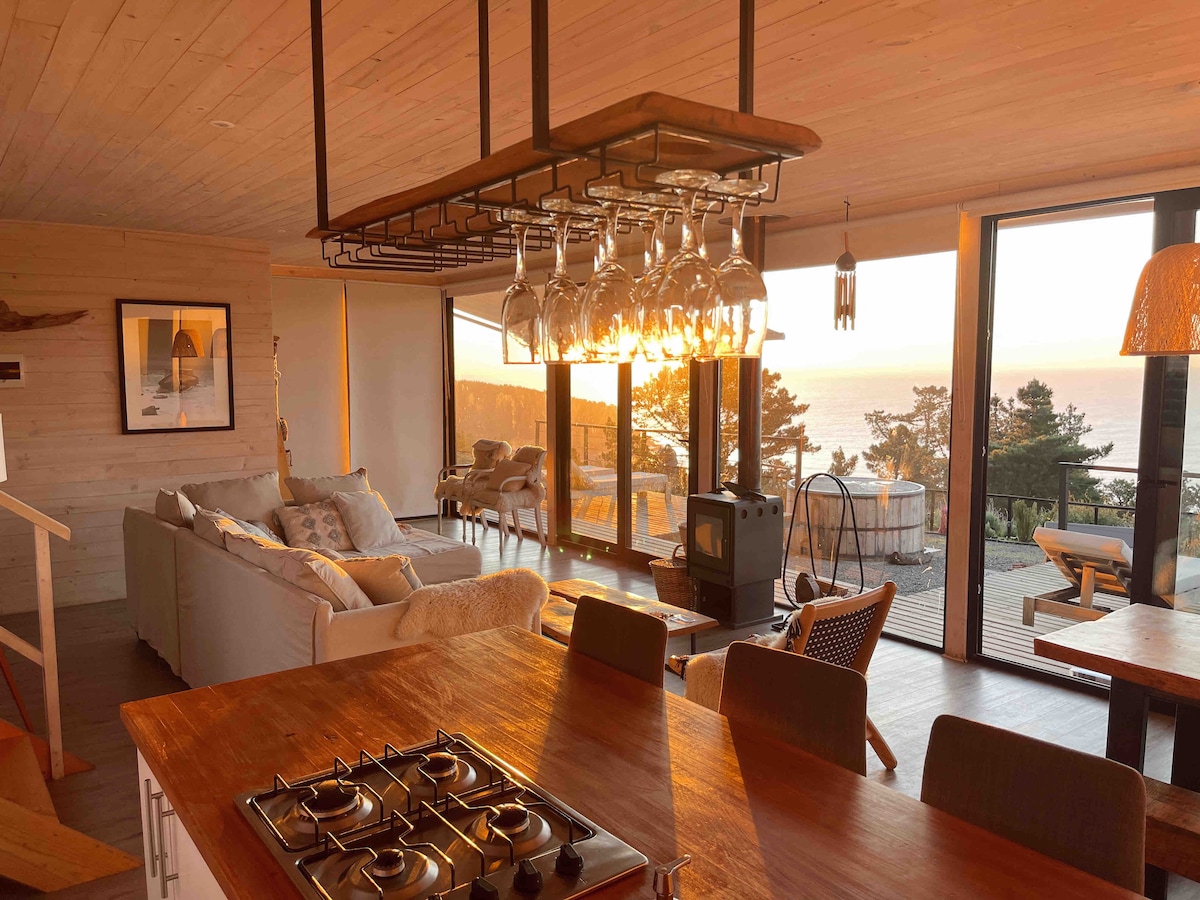 A spacious living area features large glass windows providing a view of the ocean at sunset. A contemporary dining area with a hanging light fixture showcases glasses arranged below. A cozy seating arrangement includes a sofa and armchairs, with a small wood-burning stove nearby.