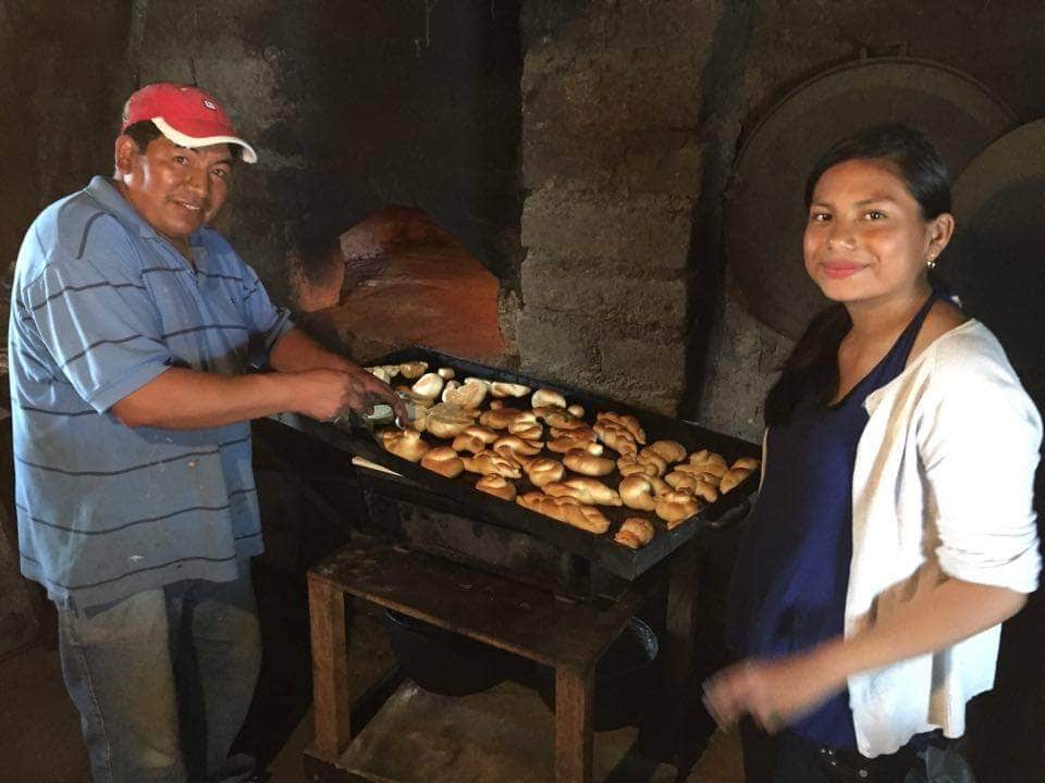 A traditional cooking scene is depicted with a large tray of baked goods placed on a rustic table. Two individuals work together inside a stone oven setting, characterized by dark tones and a warm atmosphere. The oven's opening reveals glowing embers.