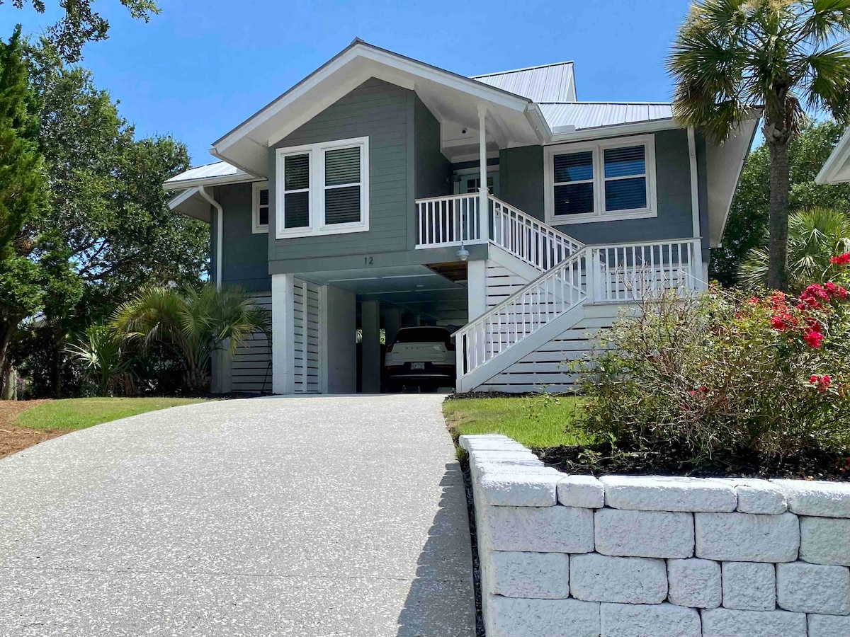 A two-story beach cottage is depicted, featuring a light grey exterior with white trim. An inviting driveway leads to the entrance, framed by lush greenery and blooming flowers. A spacious porch with white railings is visible above the carport, enhancing outdoor accessibility.