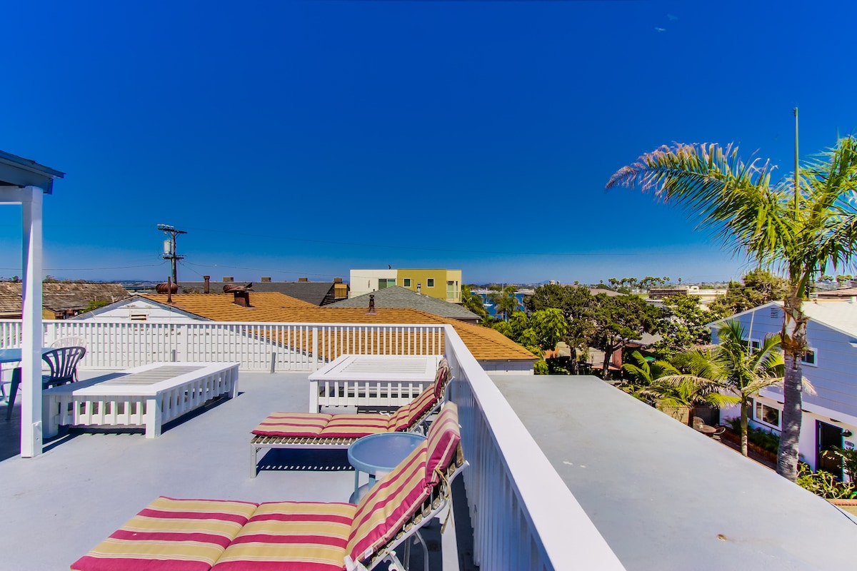 A rooftop deck offers expansive views of the neighborhood and bay. Outdoor furniture includes comfortable lounge chairs with striped cushions and a small side table. Clear skies and distant palm trees create a relaxed atmosphere, enhancing the outdoor experience.