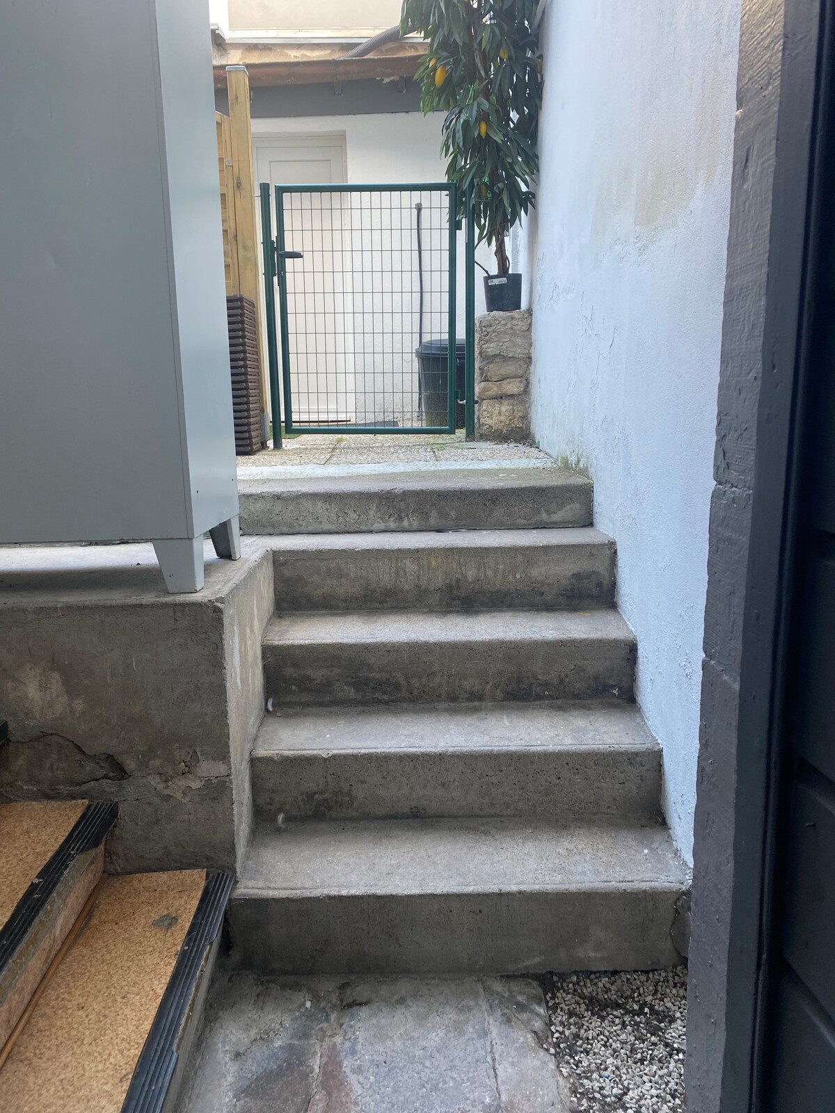 A stone pathway leads up a few steps to a secure gate framed by greenery. The surrounding walls are painted white, providing a clean and simple backdrop. The area appears tidy, with gravel and paving stones accentuating the entrance.