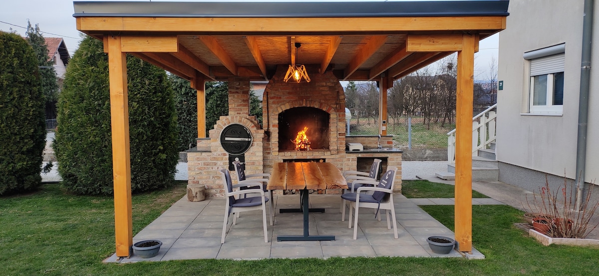 An outdoor dining area is presented under a wooden pergola, featuring a stone barbeque and a large table surrounded by four chairs. A warm fire flickers in the barbeque, and the grassy courtyard offers ample space for relaxation.