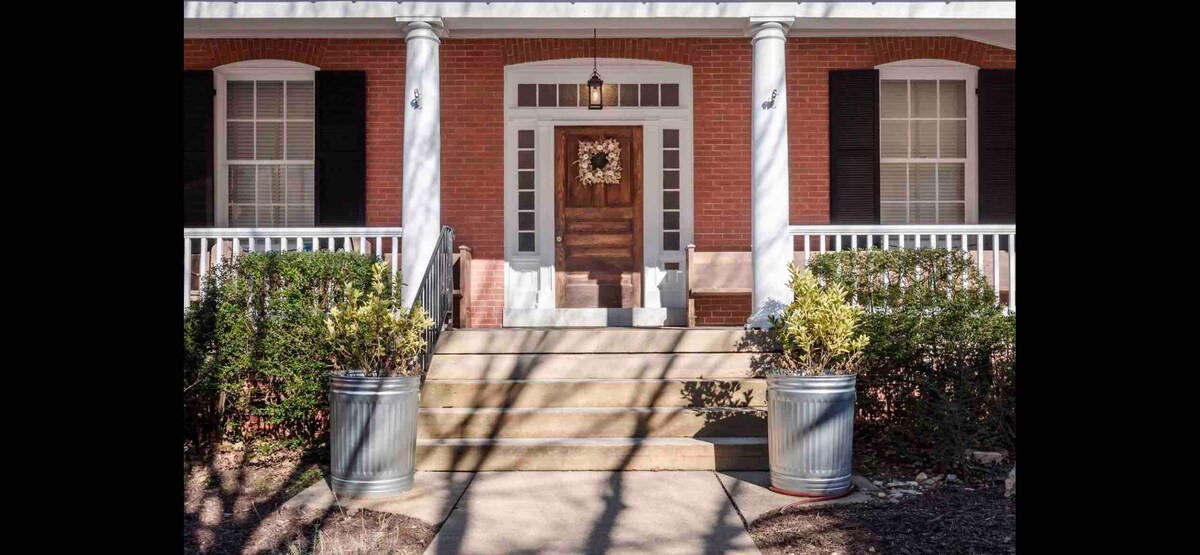 The entrance features a classic front door surrounded by large, symmetrical windows. Steps lead up to the porch, where light fixtures hang above. Potted plants line the sides, set against a backdrop of brick and white columns, creating a welcoming entryway.