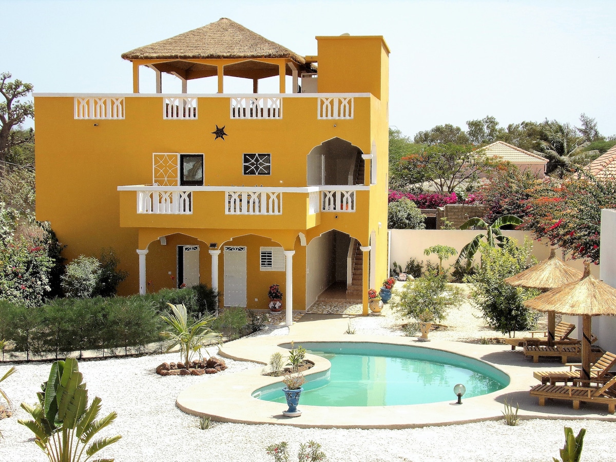 A brightly colored two-story house features a thatched roof and multiple balconies. A private pool is surrounded by landscaped gardens, with shaded seating areas and tropical plants. The exterior is accented by vibrant flowers, enhancing the inviting outdoor space.