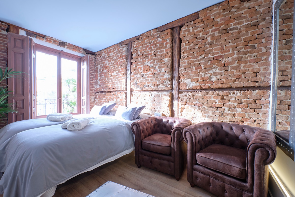 A bright room features exposed brick walls and large windows allowing natural light. A spacious bed with soft linens and two tufted armchairs provide comfortable seating. The wooden floor adds warmth to the setting, creating a cozy environment for relaxation.