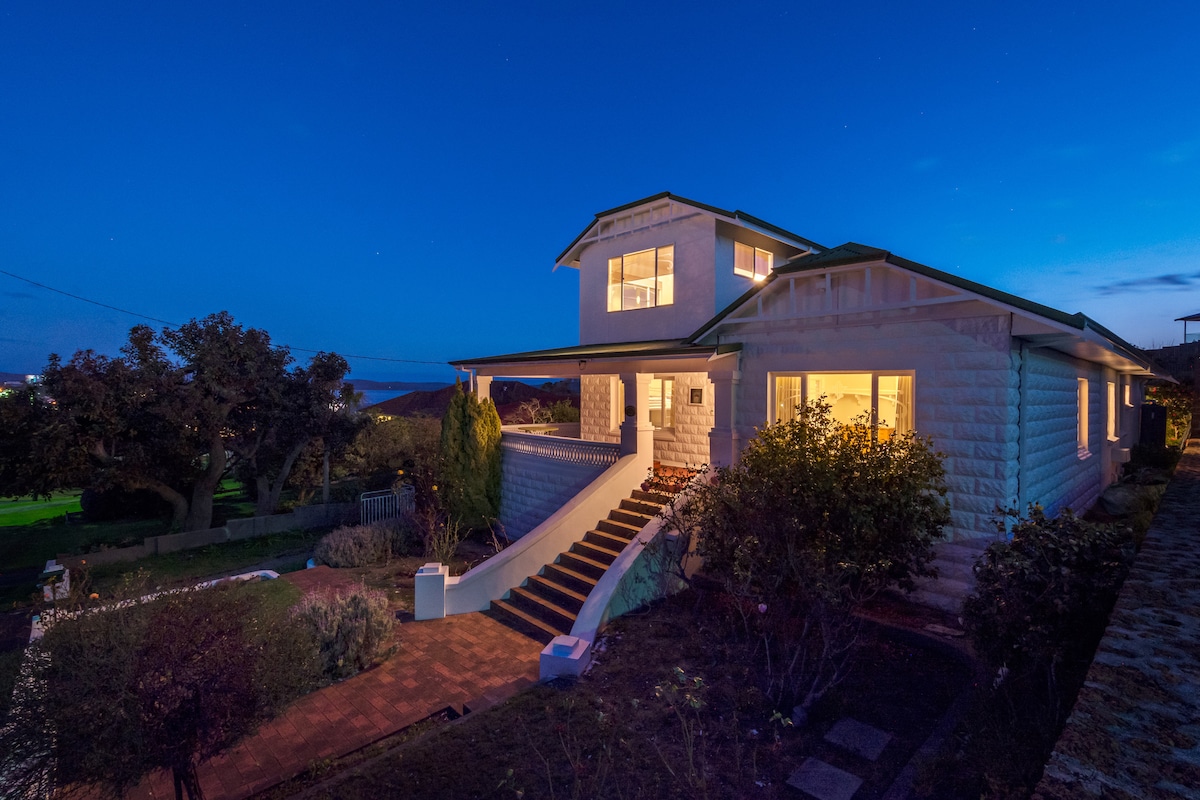 A spacious house is illuminated against a dusk sky, highlighting its inviting entrance and staircase. Surrounding greenery adds to the tranquil setting, while large windows provide a glimpse of the warm interior. The structure's unique architectural details are gently emphasized by the evening light.