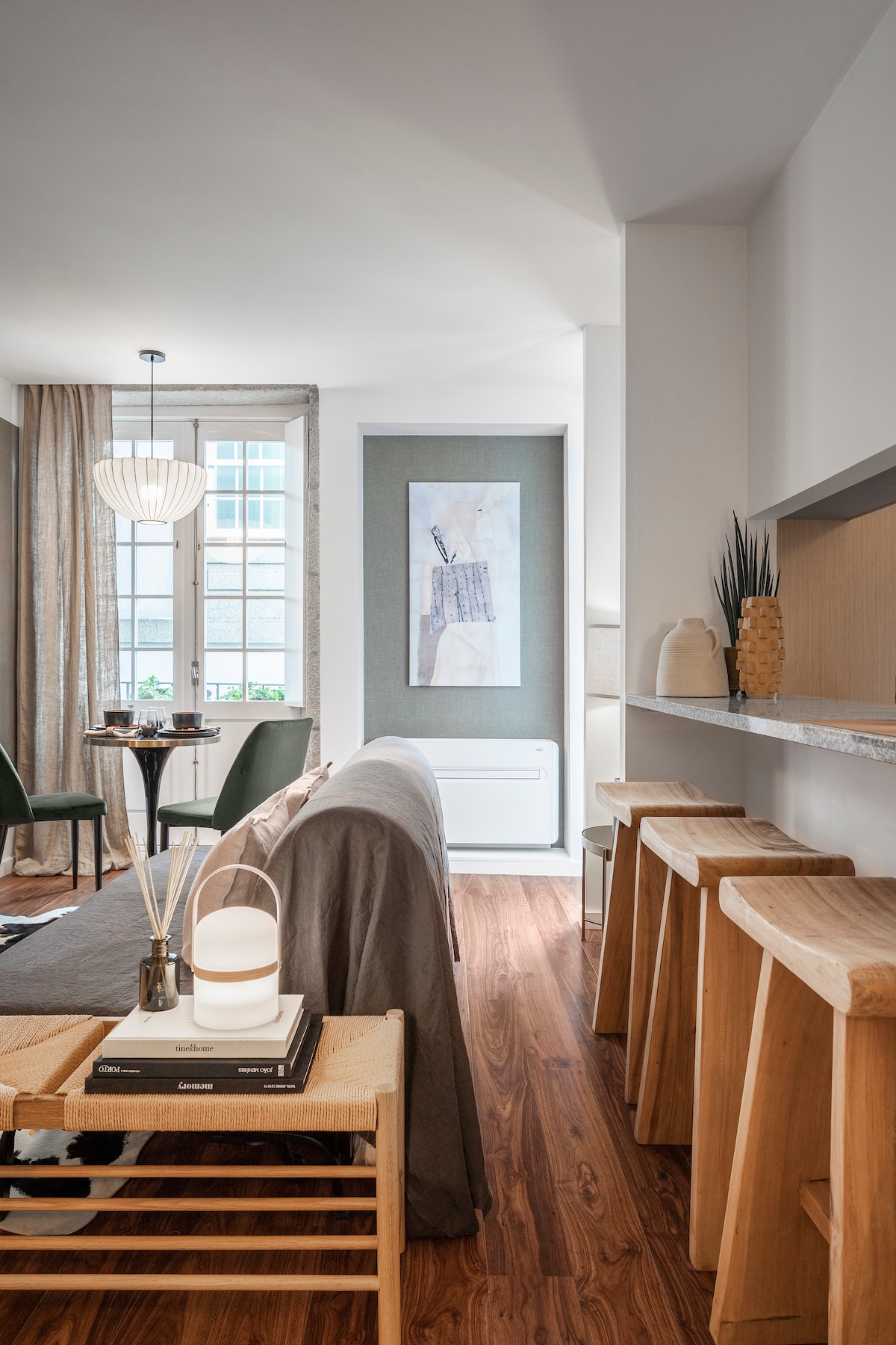 A stylish living area features a comfortable sofa with a draped throw and modern accent chairs. Natural light filters through a large window, illuminating wooden flooring. Elegant decor, including a wall art piece and decorative objects, enhances the inviting atmosphere. A spacious kitchen counter with wooden stools is visible.