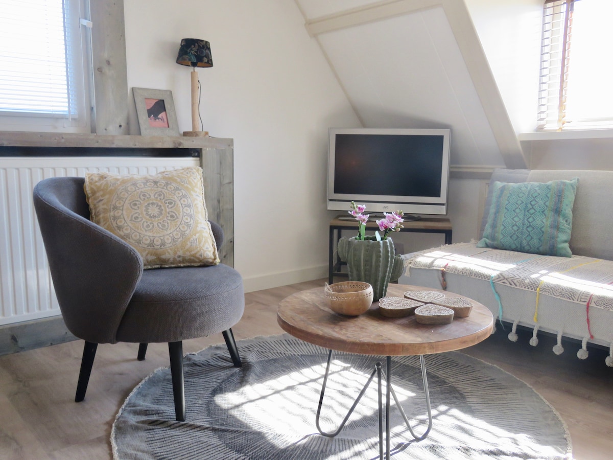 A comfortable seating area is featured, including a gray armchair with decorative pillow and a spacious sofa with patterned throw. A round wooden coffee table is placed in the center, adorned with small bowls and a potted plant, while natural light filters through nearby windows.