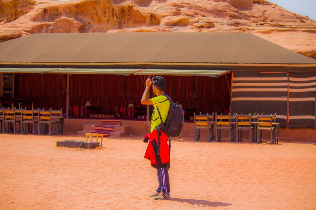 A guest stands on the sand, facing a large Bedouin-style tent. The tent features striped fabric, with wooden seating visible in front. The surrounding rocky landscape adds a natural backdrop, highlighting the desert environment.