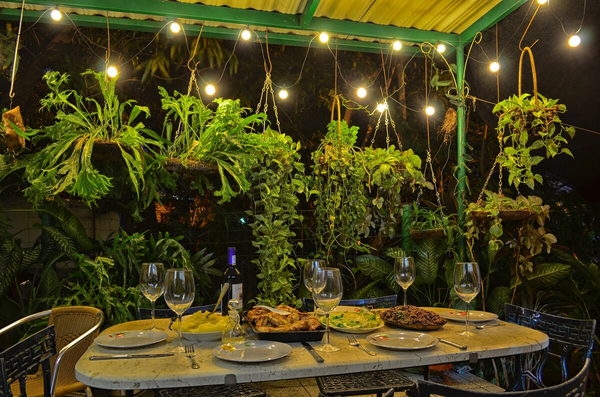 An outdoor dining area is styled with a wooden table set for a meal, featuring plates and glasses. Colorful dishes are displayed prominently. Hanging plants adorn the space, complemented by string lights that create a cozy ambiance among the lush greenery.