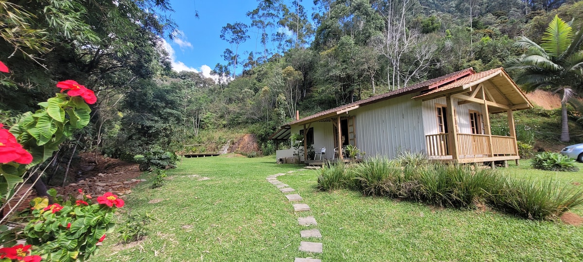 A charming house is set amidst lush greenery, with a spacious lawn in front. Pathways lead to the entrance, which is adorned with flowering plants. Surrounding trees provide a serene backdrop, creating a tranquil atmosphere perfect for relaxation.