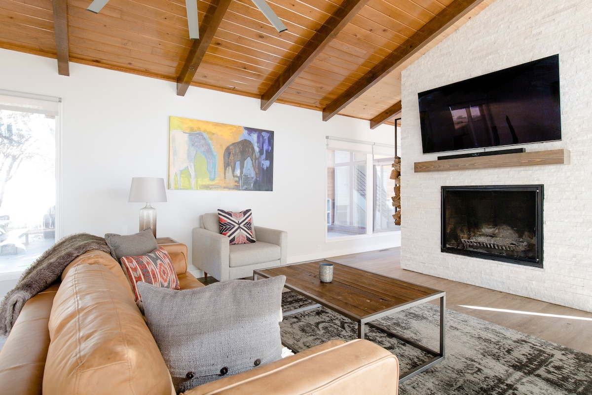 A spacious living area features a beige leather sofa and a light gray armchair, complemented by a decorative throw and patterned cushions. A large wall-mounted television is positioned above a modern fireplace, while a vibrant artwork depicts elephants. Large windows allow natural light to fill the space.