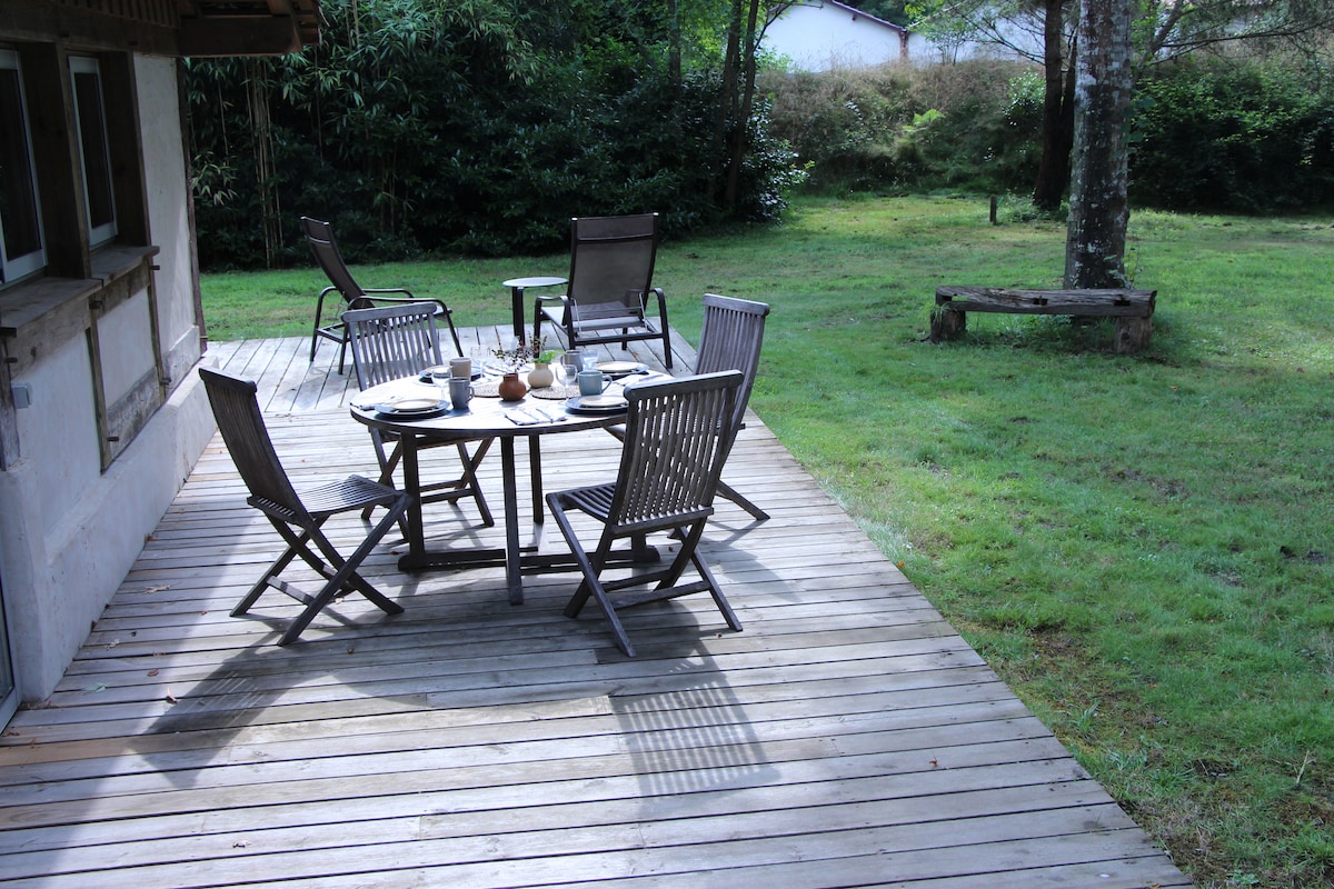A wooden deck is seen, furnished with a circular table surrounded by six folding chairs. Plates and cutlery are set on the table, suggesting a place for dining. Lush greenery surrounds the area, providing a serene natural backdrop.