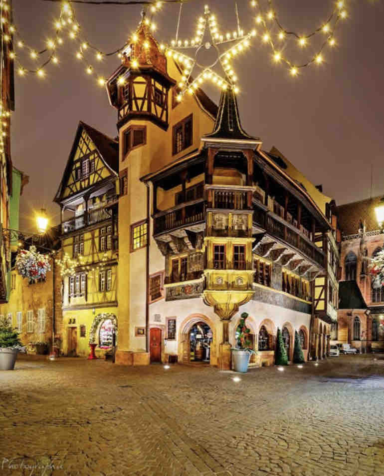 A charming building in the heart of a village is illuminated by decorative lights and a large star. The unique architecture features wooden balconies and intricate details, surrounded by cobblestone streets that create an inviting atmosphere.