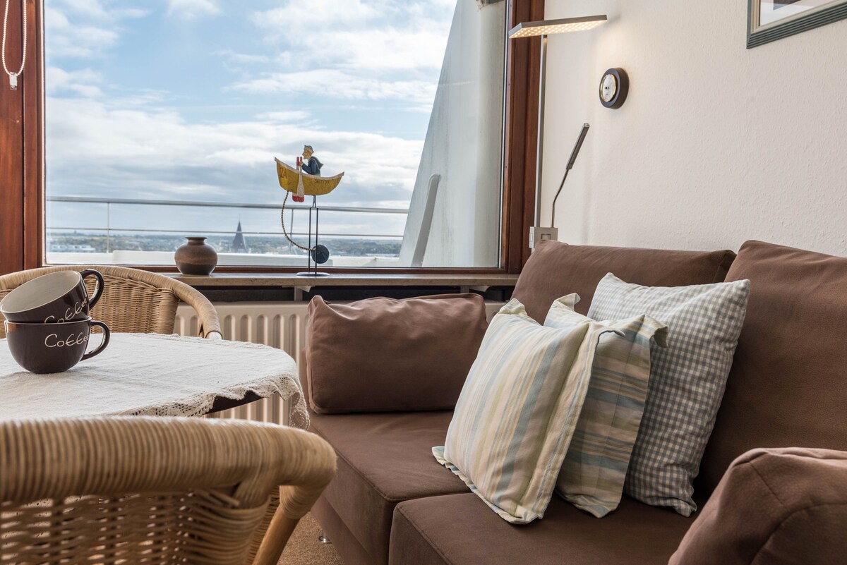 A cozy living area features a brown sofa adorned with a variety of patterned throw pillows. A round table sits nearby, accompanied by a woven chair. Natural light floods through large windows, offering a view of the surroundings.