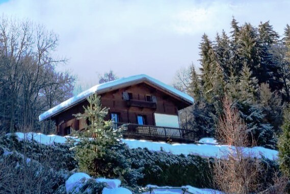 The chalet is situated on a hillside, surrounded by snow-covered trees. A wooden balcony extends from the structure, offering scenic views of the winter landscape. The chalet's roof is blanketed in fresh snow, creating a cozy alpine aesthetic.