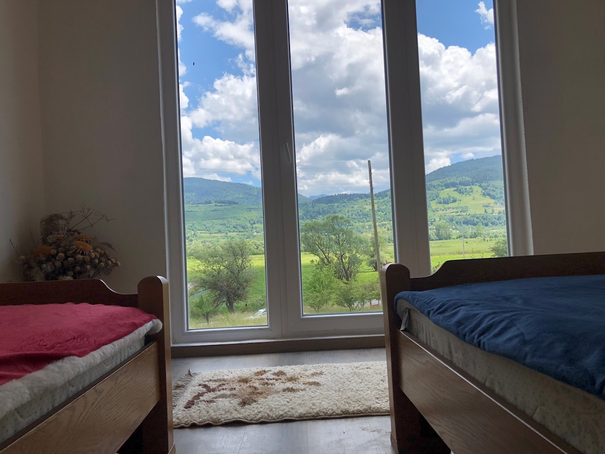 A bright room features two beds with simple linens, positioned near large glass doors. A scenic view of rolling green hills and a partly cloudy sky is framed by the doors, while a small rug adds a touch of comfort to the wooden floor.