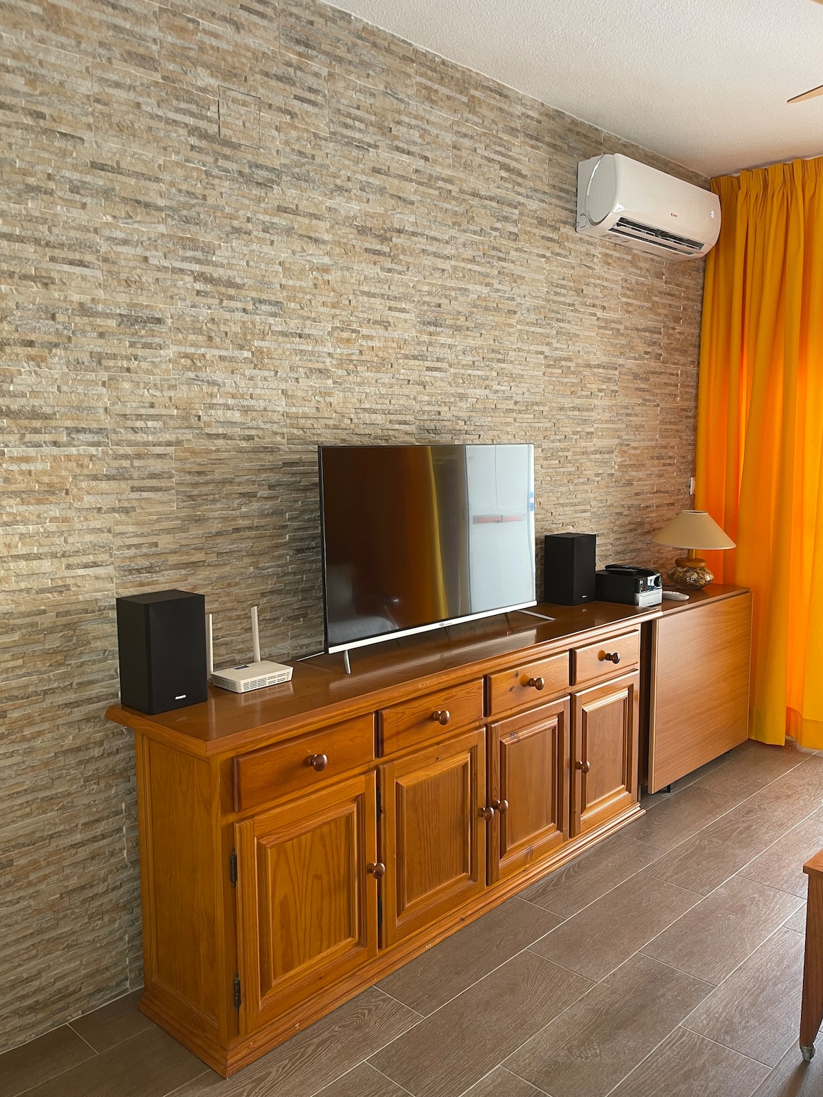 A wooden sideboard displays a television and audio speakers against a textured stone wall. Warm orange curtains frame the space, while a lamp adds subtle lighting. The flooring is wood-like, contributing to a cohesive, inviting environment.